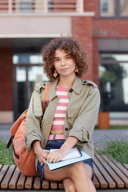 Zdjęcie portret uroczej młodej kobiety z kręconymi włosami z plecakiem jako studentka siedząca na świeżym powietrzu na kampusie uczelni uśmiechając się do kamery