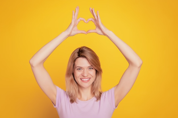 Portret uroczej słodkiej damy demonstruje symbol serca podnosi ręce na żółtym tle