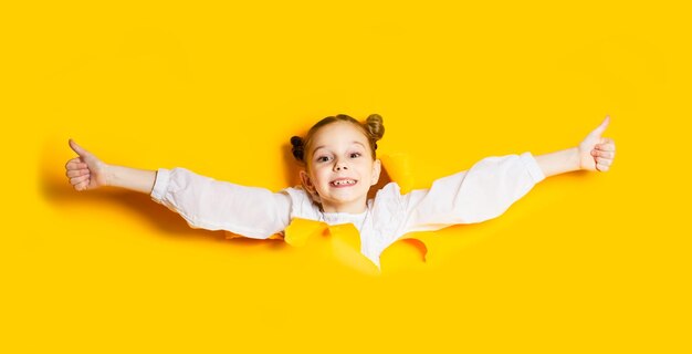 Zdjęcie portret uroczej uczennicy, która czuje się swobodnie przedzierając się przez rozdartą ścianę z żółtego papieru emocjonalna dziewczyna z radością na twarzy trzymająca kciuki w górę nagłe pojawienie się