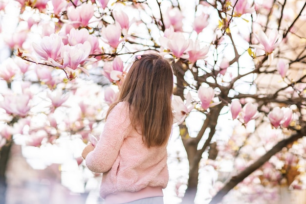 Portret uroczej wesołej dziewczyny w szarej sukience w kurtce rosa, przebywającej wiosną w parku pod kwitnącą różową magnolią.