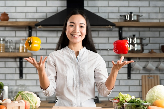 Zdjęcie portret uśmiecha się młoda kobieta żongluje z dzwonkowymi pieprzami w kuchni