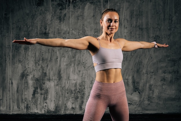 Portret uśmiechający się mięśni młoda kobieta lekkoatletycznego z doskonałe piękne ciało w odzieży sportowej robi rozciąganie, ćwiczenia rozłożone ręce i patrząc na kamery. Kaukaska kobieta fitness pozuje w studio
