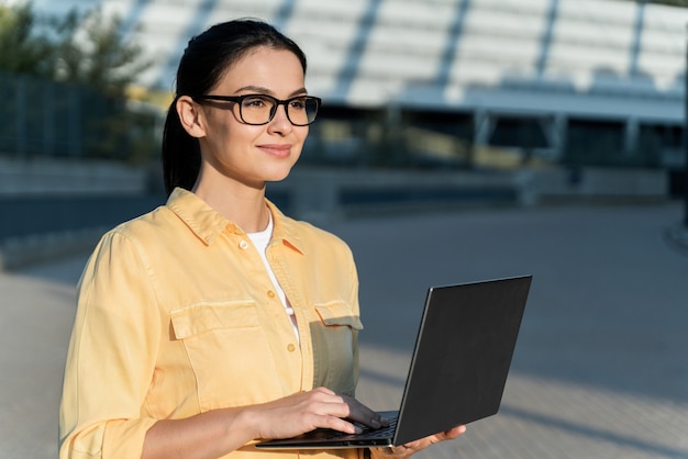 Portret uśmiechający się niezależny pracownik. Szczęśliwa dziewczyna w codziennej koszuli trzyma laptopa, używa komputera do edukacji lub pracy online, przegląda internet na ulicy