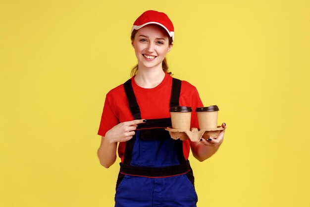 Portret uśmiechający się pozytywny kurier kobieta stojąca i wskazująca na filiżanki kawy, patrząc na kamery z uśmiechem na sobie kombinezon i czerwoną czapkę Kryty studio strzał na białym tle na żółtym tle