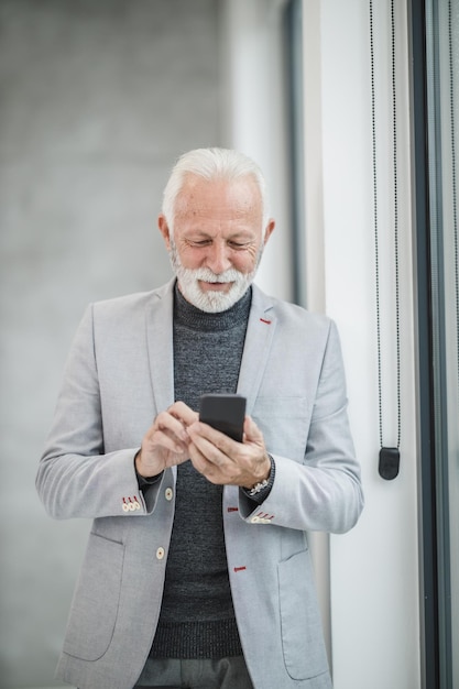 Portret uśmiechający się starszy biznesmen za pomocą aplikacji na smartfonie stojąc w nowoczesnym biurze.