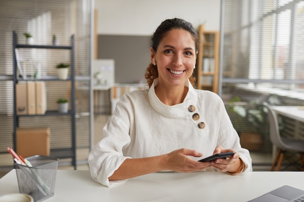 Portret uśmiechnięta bizneswoman trzymając smartfon i patrząc na kamery siedząc przy biurku w biurze, kopia przestrzeń