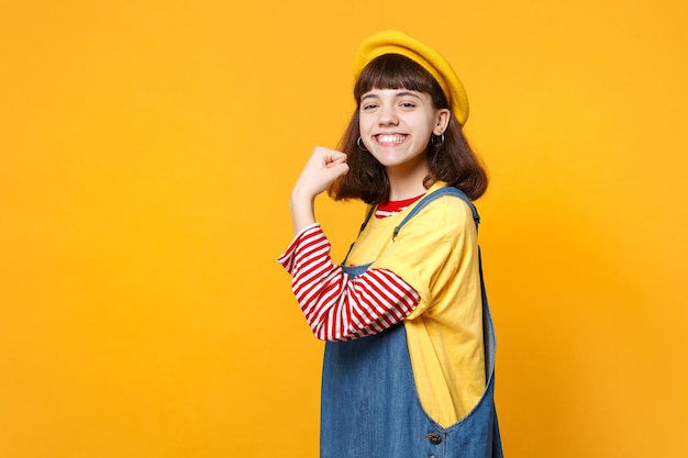 Portret uśmiechnięta dziewczyna nastolatka w francuski beret, dżinsowa sukienka pokazując biceps, mięśnie na białym tle na tle żółtej ściany w studio. Ludzie szczere emocje, koncepcja stylu życia. Makieta miejsca na kopię.