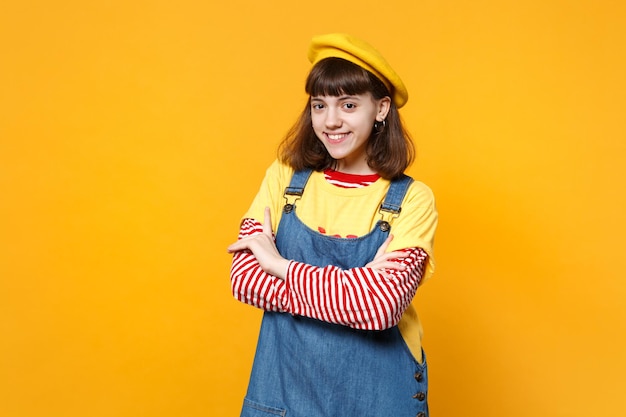 Portret uśmiechnięta dziewczyna nastolatka w francuski beret i denim sundress trzymając ręce skrzyżowane na białym tle na tle żółtej ściany w studio. Ludzie szczere emocje, koncepcja stylu życia. Makieta miejsca na kopię.