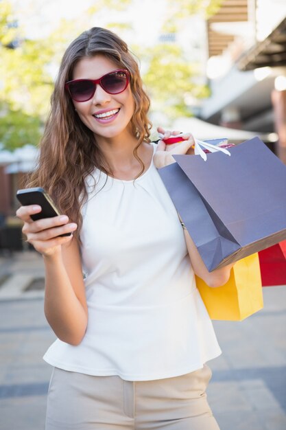 Portret uśmiechnięta kobieta z okularami przeciwsłonecznymi i torba na zakupy używać jej smartphone