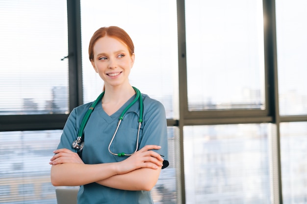 Portret uśmiechnięta lekarka w niebieskim zielonym mundurze stojącym rękami skrzyżowanymi w pobliżu okna w słoneczny dzień w gabinecie przychodni medycznej.