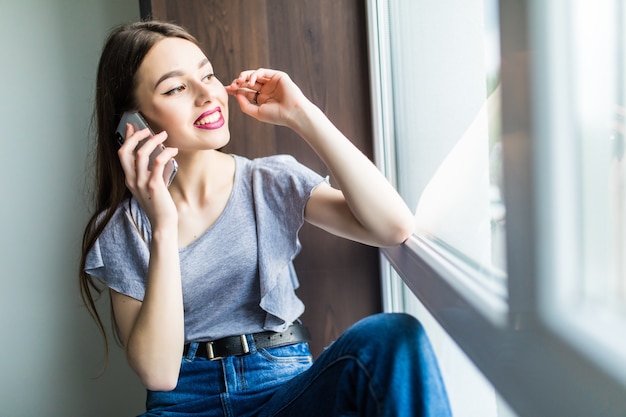 Portret Uśmiechnięta Młoda Dorosła Kobieta Siedzi Na Parapecie Z Laptopem, Rozmawiając Przez Telefon Komórkowy, Odwraca Wzrok.