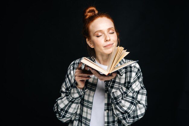 Zdjęcie portret uśmiechnięta młoda kobieta studentka stojąca z zamkniętymi oczami, trzymająca otwartą książkę na na białym tle czarnym tle ładna rudowłosa modelka emocjonalnie pokazująca mimikę w studio