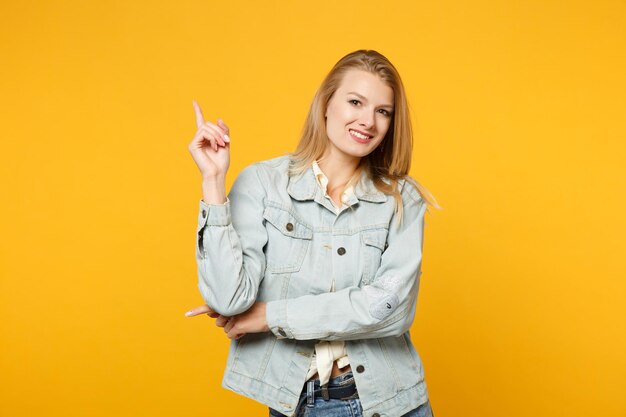 Portret uśmiechnięta młoda kobieta w dżinsowych ubraniach casual, wskazując palcem w górę, patrząc na kamery na białym tle na żółto pomarańczowe ściany w studio. Koncepcja życia ludzi. Makieta miejsca na kopię.