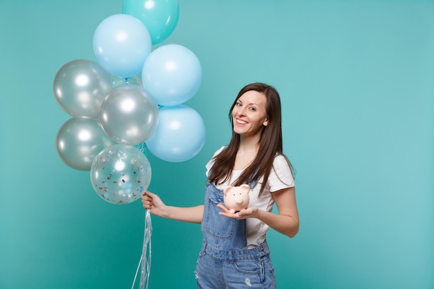 Portret uśmiechnięta młoda kobieta w dżinsowych ubraniach trzymając skarbonkę z okazji z kolorowych balonów na białym tle na niebieskim tle turkus. Urodziny wakacje, koncepcja emocje ludzi.