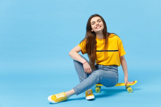 Portret uśmiechnięta młoda kobieta w żywych ubraniach casual, patrząc na kamery i siedząc z żółtą deskorolką na białym tle na tle niebieskiej ściany w studio. Koncepcja życia ludzi. Makieta miejsca na kopię.
