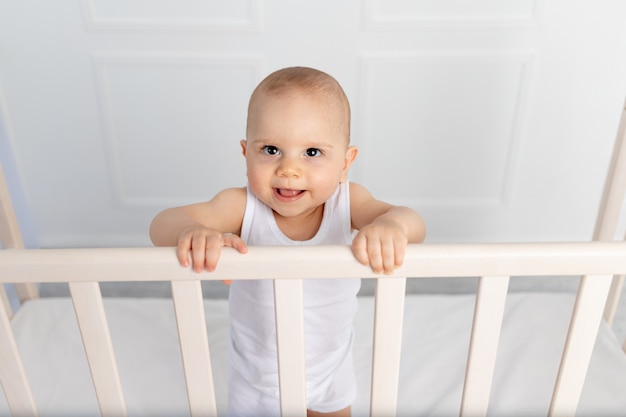 Portret Uśmiechniętego Chłopca W Wieku 8 Miesięcy Stojącego W łóżeczku W Pokoju Dziecięcym W Białych Ubraniach Il, Poranne Dziecko, Koncepcja Produktów Dla Dzieci