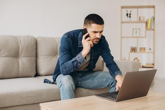 Portret uśmiechniętego faceta rozmawiającego przez telefon komórkowy, pracującego na laptopie, siedzącego na kanapie w saloniexDxA