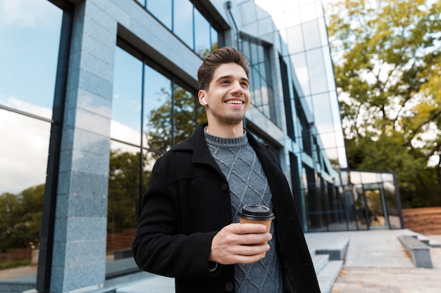 Portret uśmiechniętego mężczyzny w wieku 30 lat w nausznikach trzymającego papierowy kubek kawy na wynos podczas spaceru przed szklanym budynkiem