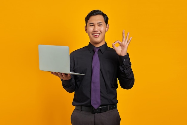 Portret uśmiechniętego młodego azjatyckiego biznesmena pokazującego znak porządku i trzymającego laptopa