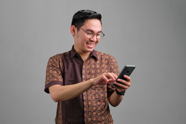 Portret uśmiechniętego młodego azjatyckiego mężczyzny w batikowej koszuli za pomocą smartfona do przesyłania wiadomości z przyjaciółmi na białym tle na szarym tle