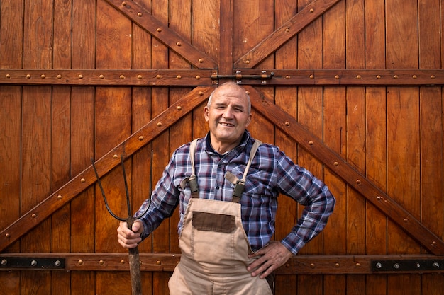 Portret Uśmiechniętego Pracowitego Rolnika Posiadającego Widły Narzędzie I Stojącego Przy Drzwiach Drewnianej Stodoły Lub Spichlerza żywności W Gospodarstwie Zwierząt Domowych.