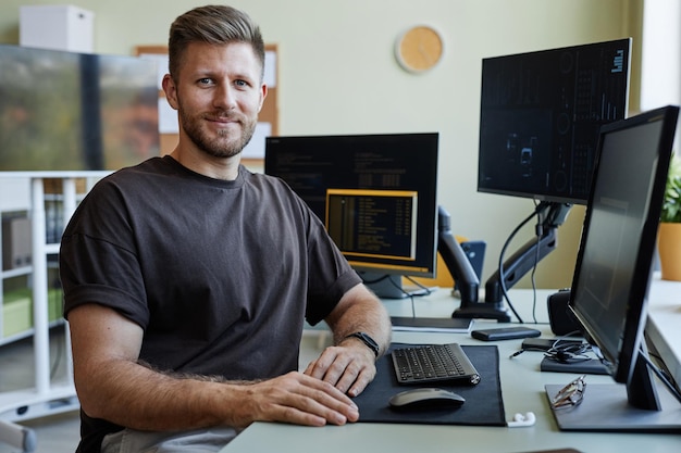 Portret uśmiechniętego programisty patrzącego na kamerę siedząc przy biurku z komputerami
