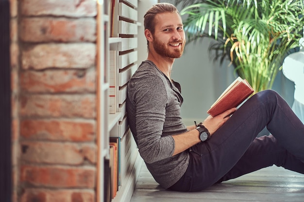 Portret uśmiechniętego, przystojnego, stylowego rudego studenta w zwykłych ubraniach, siedzącego na podłodze w domu i trzymającego książkę, patrzącego w kamerę.