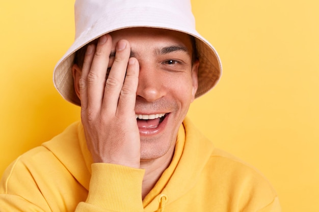 Portret Uśmiechniętego Szczęśliwego Młodego Dorosłego Mężczyzny Noszącego Bluzę Z Kapturem W Stylu Casual I Pozowanie Panama Na Białym Tle Nad żółtym Tłem Zakrywającym Połowę Twarzy I Oczu Dłonią