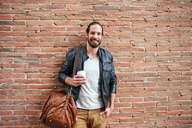 Portret uśmiechniętego zadowolonego Europejczyka z wiązanymi włosami, patrzącego na ciebie, stojącego ze skórzaną torbą nad murem na ulicy miasta i pijącego kawę na wynos