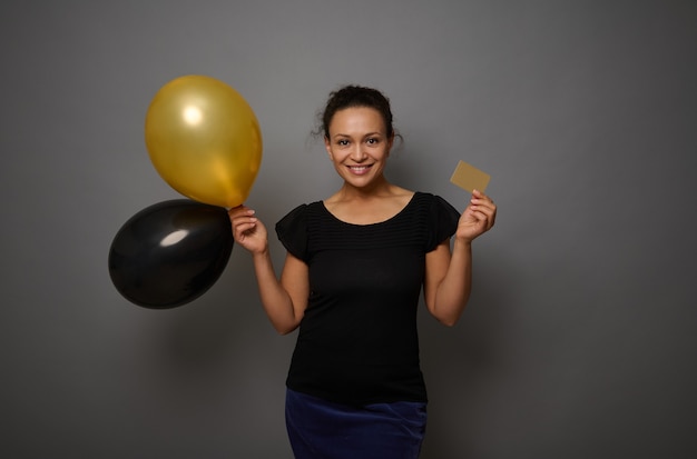 Portret uśmiechniętej afrykańskiej kobiety trzymającej złotą kartę kredytową lub rabatową i napompowane czarno-złote balony i uśmiechy patrząc na kamerę pozującą na szarym tle ściany z kopią przestrzeni reklamowej