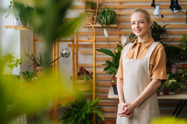 Portret uśmiechniętej atrakcyjnej kobiecej kwiaciarni w fartuchu stojącej pozowanie w kwiaciarni patrząc na kamerę