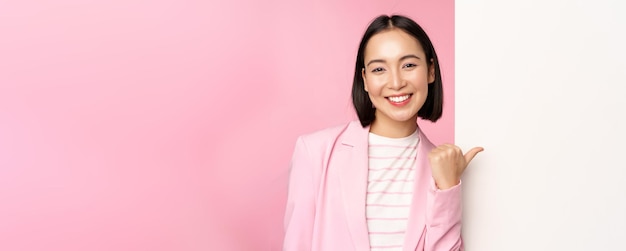 Portret uśmiechniętej azjatyckiej bizneswoman w garniturze korporacyjnej damy, wskazując palcem na białą pustą tablicę ścienną z informacją lub reklamą stojącą na różowym tle