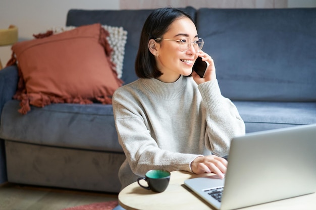 Portret uśmiechniętej azjatyckiej kobiety wykonującej telefon, pracującej na laptopie i rozmawiającej na smart