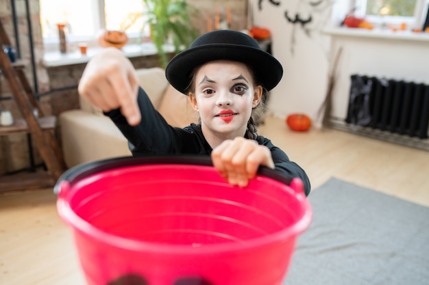 Zdjęcie portret uśmiechniętej dziewczyny mim z sercem narysowanym na ustach wskazujących na wiadro i proszących o włożenie cukierków na halloween