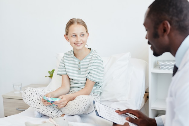 Portret uśmiechniętej dziewczyny rozmawiającej z lekarzem siedząc na łóżku w sali szpitalnej, kopia przestrzeń