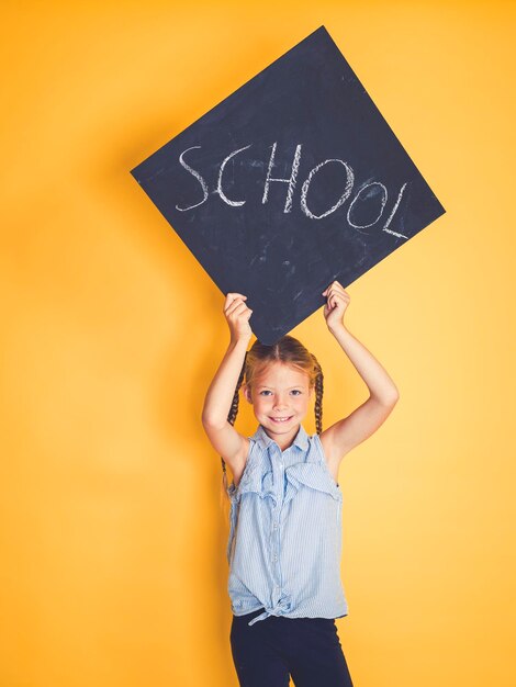 Zdjęcie portret uśmiechniętej dziewczyny trzymającej tablicę z szkolnym tekstem, stojącej na żółtym tle