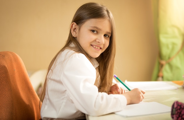 Portret uśmiechniętej dziewczyny w białej koszuli siedzącej za biurkiem i odrabiającej pracę domową