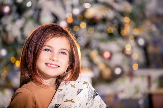 Portret uśmiechniętej dziewczyny z chytrym planem przeciwko choince z prezentem w dłoniach.