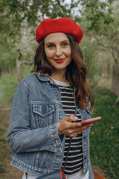 Portret uśmiechniętej kobiety w berecie z czerwonymi ustami za pomocą smartfona i patrząc na kamerę w parku