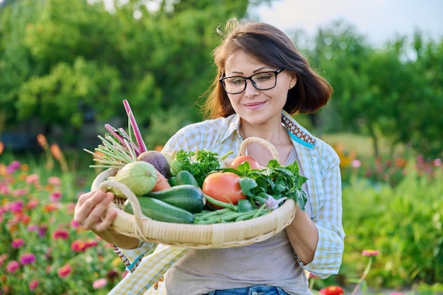 Portret uśmiechniętej kobiety w średnim wieku z koszem różnych świeżych surowych warzyw i ziół lato natura ogród warzywny tło Zbiory z gospodarstwa ekologicznego zdrowa żywność odżywianie ogrodnictwo