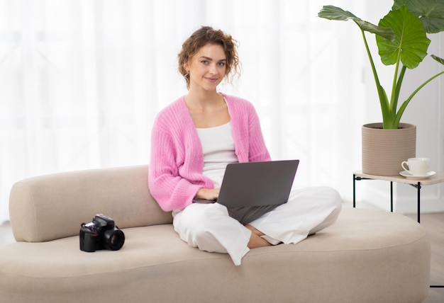Portret uśmiechniętej kobiety z laptopem siedzącej na kanapie obok aparatu fotograficznego
