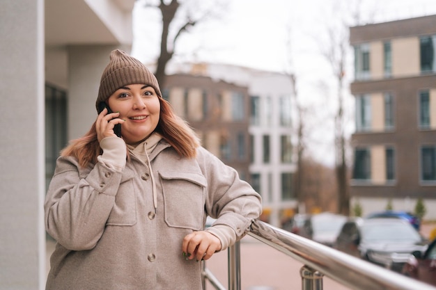 Portret uśmiechniętej kobiety z nadwagą w ciepłym kapeluszu i kurtce rozmawiającej na telefonie komórkowym stojącej w pobliżu poręczy na ulicy miasta w pochmurny jesienny dzień