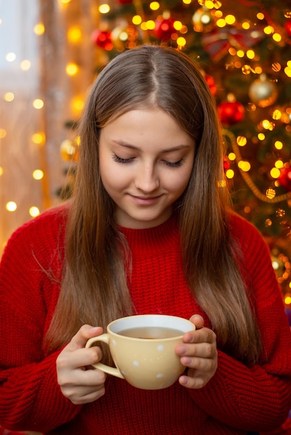 Portret uśmiechniętej młodej blond dziewczyny w ciepłym czerwonym swetrze trzymającym filiżankę herbaty w dłoniach