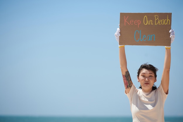 Portret uśmiechniętej młodej kobiety stojącej na plaży z ręcznie robionym plakatem utrzymuj naszą plażę w czystości