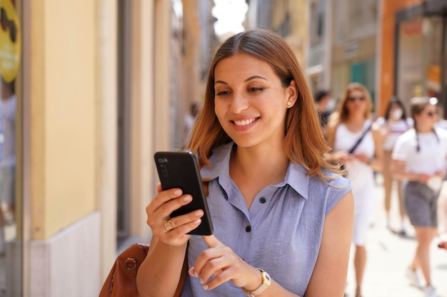 Portret uśmiechniętej młodej kobiety używającej smartfona na ulicy z ludźmi chodzącymi w tle