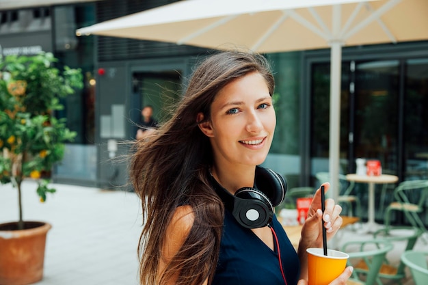 Portret uśmiechniętej młodej kobiety ze słuchawkami i napojem na wynos w mieście