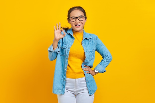 Portret uśmiechniętej młodej studentki azjatyckiej kobiety w dżinsowych ubraniach pokazujących dobry gest pokazuje symbol aprobaty na białym tle na żółtym tle. Edukacja w koncepcji uniwersytetu uniwersyteckiego