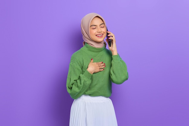Portret uśmiechniętej pięknej azjatyckiej kobiety w zielonym swetrze i hidżabie rozmawiającej na telefonie komórkowym z ręką na klatce piersiowej odizolowanej na fioletowym tle