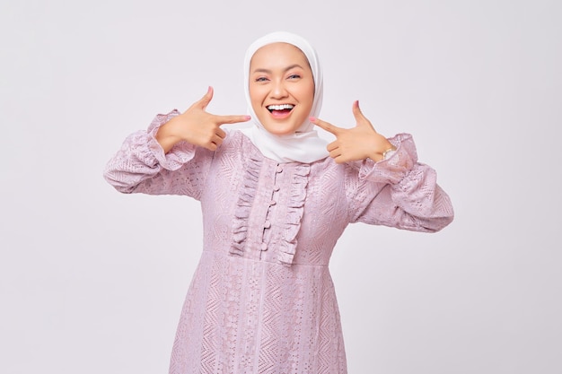 Portret uśmiechniętej pięknej młodej azjatyckiej muzułmanki noszącej hidżab i fioletową sukienkę wskazującą palcem na zęby uśmiechu na białym tle na tle białego studia