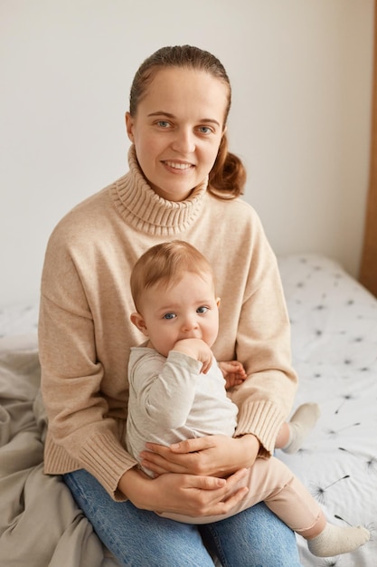 Portret uśmiechniętej pozytywnej kobiety z kucykiem, noszącej beżowy sweter z niemowlęcym dzieckiem, młodą matką siedzącą na łóżku i patrzącą na kamerę z szczęśliwym wyrazem twarzy